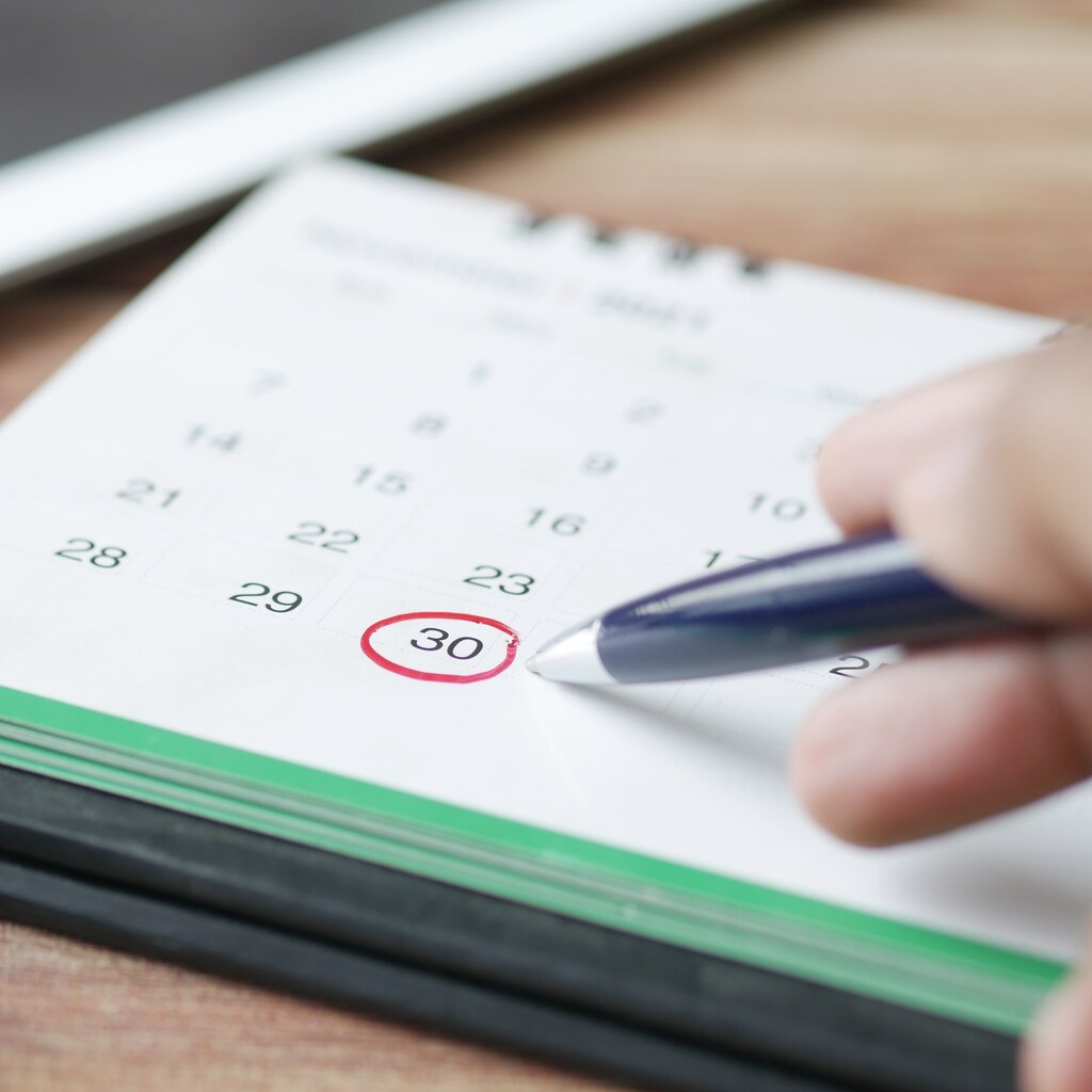 A photo of a hand marking a date on a paper calendar with a pen.