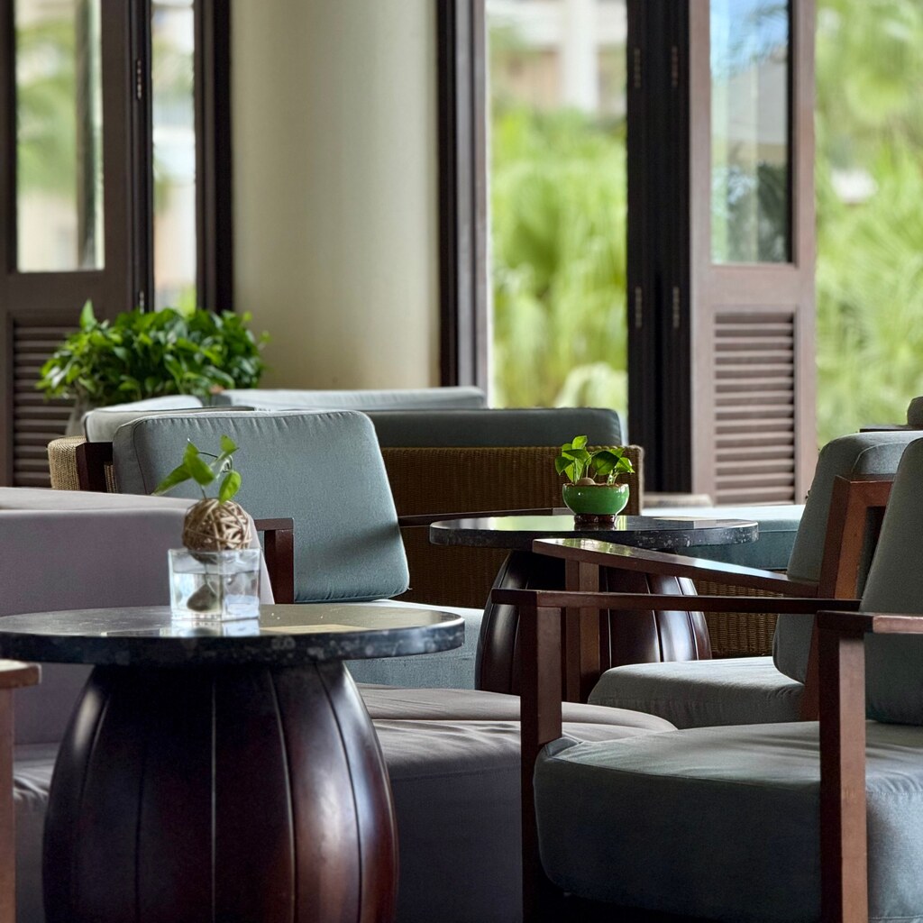 A photo of a lounge with armchairs, tables, plants, and tall windows.