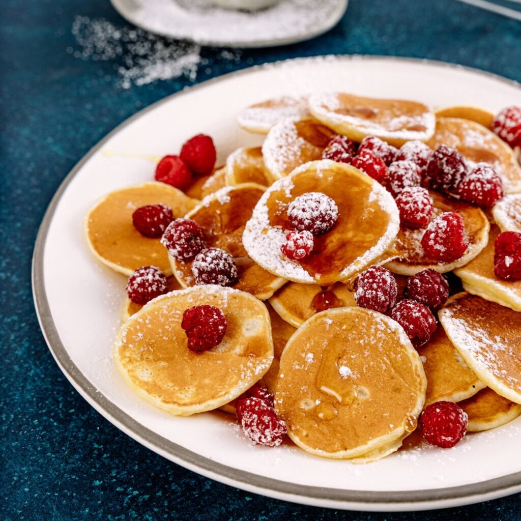 A photo of small pikelets topped with raspberries and powdered sugar.