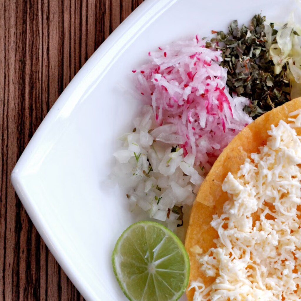A photo of a Mexican tostada topped with cheese, radish, onion, herbs, and lime.